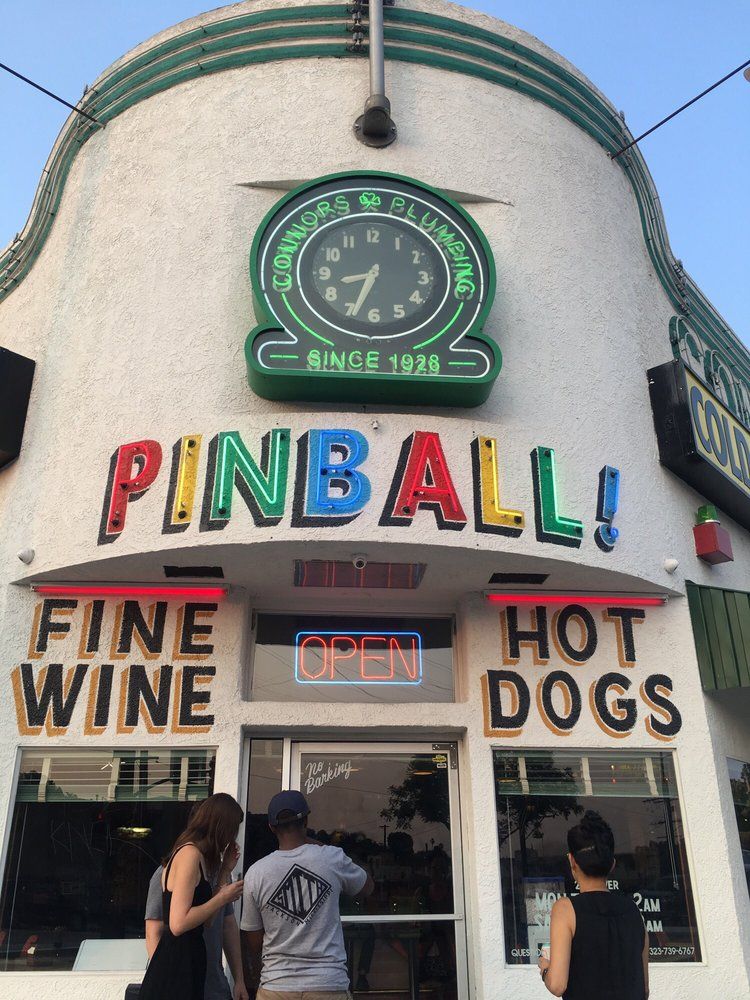 The corner of Walt's Bar at dusk: the green neon Connors Plumbing clock above hand-painted signs reading Pinball, Fine Wine and Hot Dogs, with a neon Open sign over the door