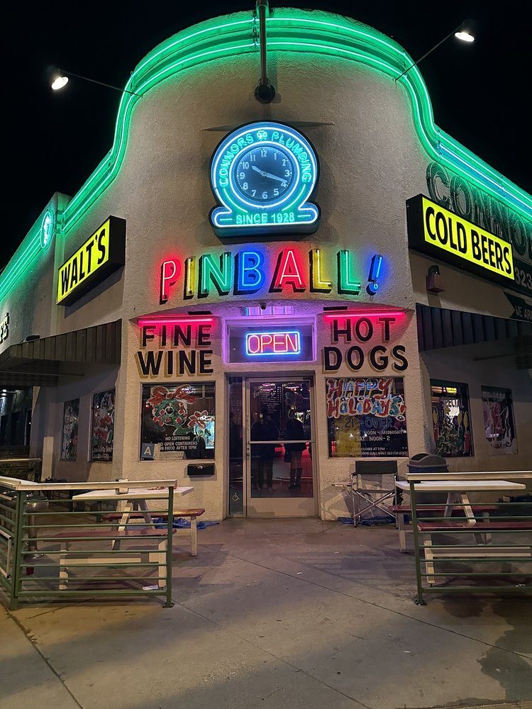 Walt's Bar at night with every sign lit: green neon around the roofline, the Connors Plumbing clock glowing above hand-painted Pinball, Fine Wine and Hot Dogs signs, and a neon Open sign over the door